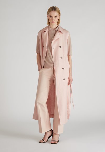 Un long gilet en lin rose avec des boutons, porté avec un haut rose clair assorti et un pantalon large. Des talons noirs à lanières complètent l'ensemble.