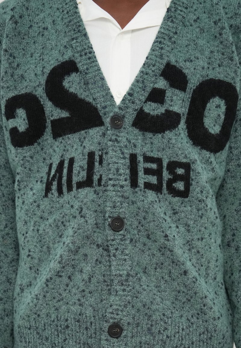 Green knitted cardigan with black lettering, v-neck, button front, and a soft, textured fabric. Notable speckled pattern throughout.