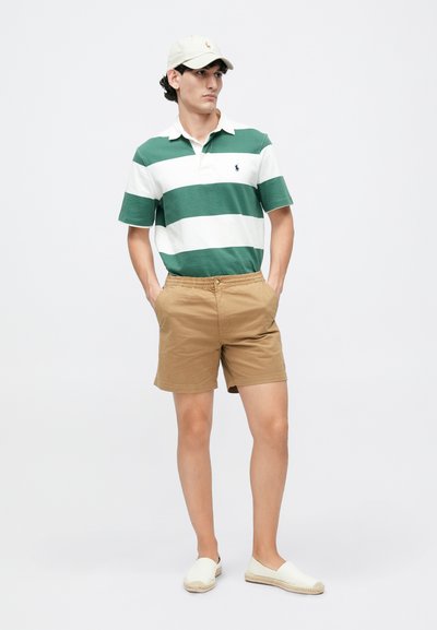 Polo din bumbac în dungi verzi și albe, pantaloni scurți kaki și pantofi slip-on într-o nuanță deschisă. Șapcă cu logo. Outfit casual de vară.