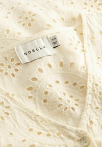 Ύφασμα σε κρεμ χρώμα με κέντημα eyelet και ετικέτα της μάρκας Noella που δείχνει το μέγεθος S/M, κατασκευασμένο στην Ιταλία, με εμφανές λευκό κουμπί.