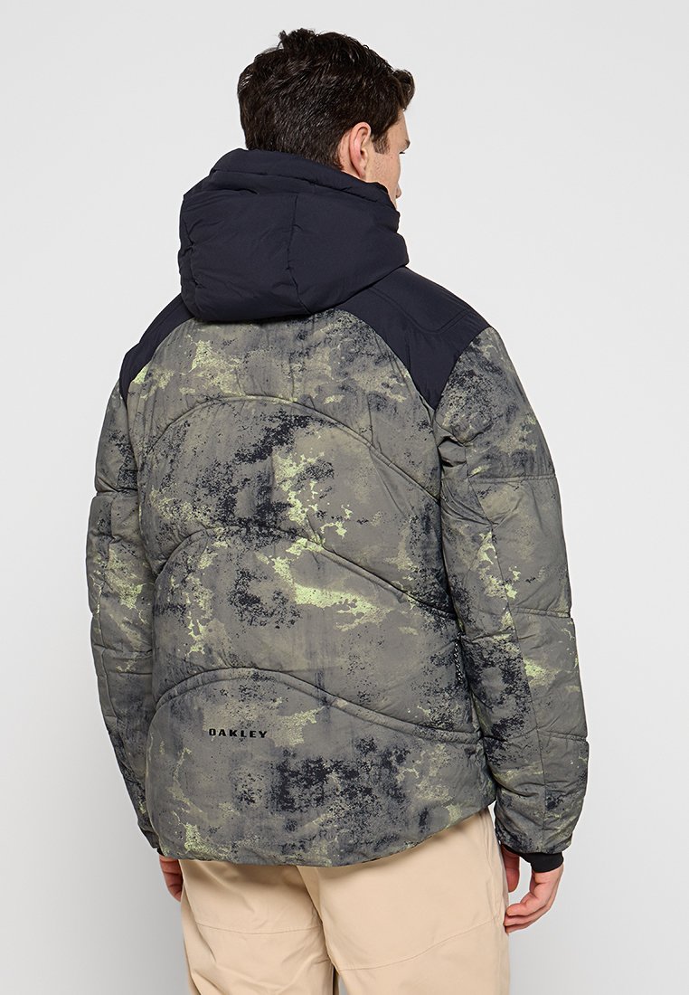 Manteau matelassé au motif camouflage avec des nuances de vert et de noir, doté d'une capuche navy et d'un texte de marque dans le bas du dos.
