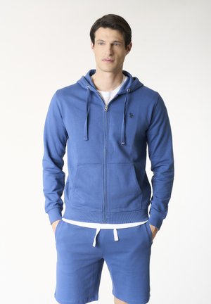 Uomo che indossa una felpa con cappuccio blu con zip e pantaloncini blu abbinati, in piedi con le mani in tasca su uno sfondo semplice.