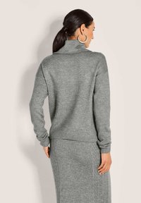 Frau mit dunklem Haar im Pferdeschwanz trägt große Creolen, einen grauen Rollkragenpullover und einen passenden Strickrock, von hinten gesehen.