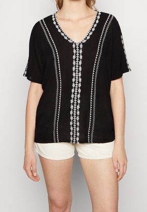 Frau trägt eine schwarze V-Ausschnitt-Bluse mit weißer vertikaler Stickerei, kombiniert mit hellbeigen Shorts, steht vor einem schlichten Hintergrund.