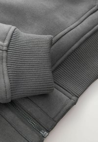 Close-up of gray sweatshirt fabric featuring ribbed cuffs and a partially visible metal zipper with "SAB" engraving.
