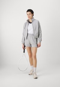 Grå idrettsjakke og shorts med elastisk liv, hvit topp, holder en tenniskølle, iført lette sneakers og sokker.