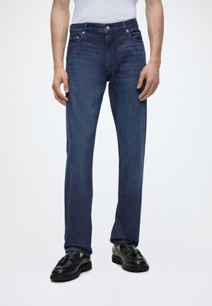 Mann trägt gerade geschnittene dunkelblaue Jeans, schwarze Tassel-Loafer und ein weißes Hemd, steht vor einem schlichten weißen Hintergrund.