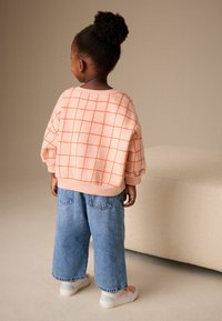 Růžová mikina s oranžovým mřížkovým vzorem; volný střih. Modré džíny s širokými nohavicemi. Světle barevné tenisky s pastelovými akcenty.