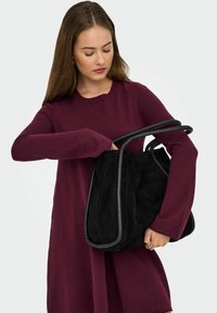 Femme dans une robe longue à manches longues bordeaux regardant dans et tenant un grand sac à main en daim noir avec des poignées en cuir enroulées.