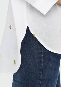 Camicia in lino bianca con ricamo di ananas giallo, orlo curvato e spacchi laterali, abbinata a jeans di denim blu scuro con una superficie liscia.