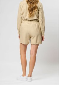 Beige linnen rompertje met een ontspannen pasvorm, elastische tailleband en opgerolde shorts. Gestyled met witte sneakers, zichtbaar vanuit de achterkant.