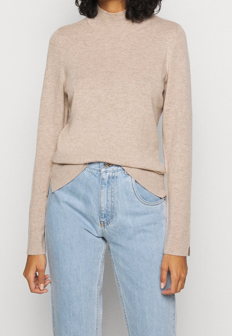 Femme portant un pull beige à manches longues avec un léger chevauchement à l'ourlet, associé à un jean taille haute bleu clair.