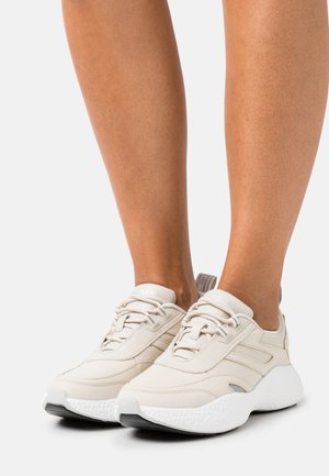 Béžové nízké tenisky s tlustou bílou podrážkou nošené na holých nohách, s šněrováním a značkovým designem Calvin Klein Jeans.