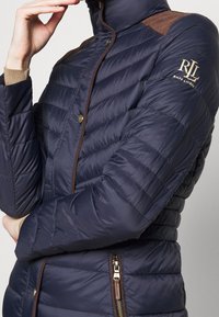 Marineblauer Pufferjacke mit horizontal gestepptem Design, braunen Schulterakzenten, goldenem Logo am Ärmel, satinierter Oberfläche und Reißverschlusstaschen.