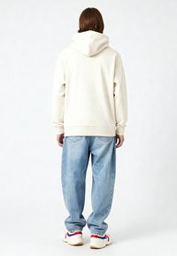 Felpa crema con un grande cappuccio nella parte posteriore, abbinata a jeans azzurri larghi e scarpe da ginnastica bianche con dettagli rossi e blu.