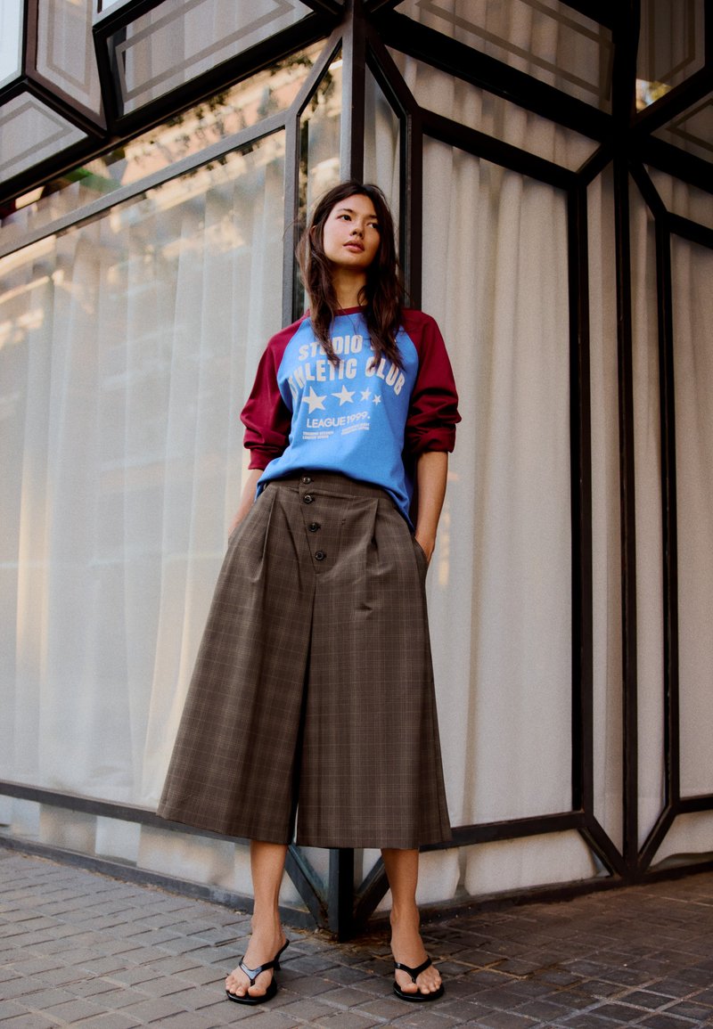 Jeune femme portant un sweat bleu et bordeaux, un large pantalon culotte à carreaux marron et des sandales noires, debout contre un angle d’un bâtiment en verre et métal.