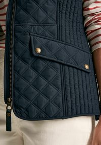 Kék steppelt mellény gyémántmintával, aranyszínű részletekkel, ferde cipzárral és alacsony, ferdén elhelyezett zsebbel, amely patenttal záródik.