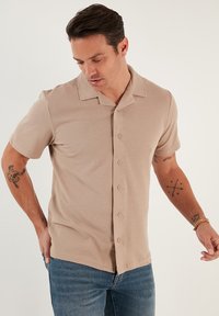 Beige kortærmet skjorte med krave i struktureret stof, med knaplukning foran og to knapper på kraven. Bæres over blå denim.