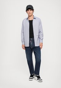Himmelblaues Hemd mit geradem Schnitt, getragen über einem schwarzen Tanktop. Kombiniert mit dunkelblauen Jeans und schwarz-weißen Sneakers.