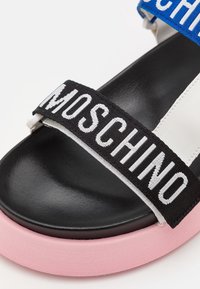 Musta sandaali vaaleanpunaisella pohjalla, jossa on valkoiset ja siniset säädettävät hihnat. Mustassa hihnassa on "MOSCHINO"-brändäys valkoisilla kirjaimilla.