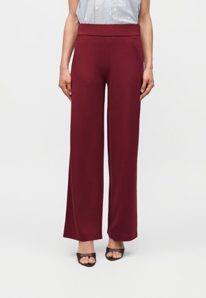 Femme portant un pantalon bordeaux taille haute à jambes larges avec poches latérales et des talons ouverts noirs, debout devant un fond uni.