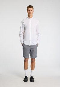 Balta pogām uz augšu veste, pelēki šorti ar sarullētām piedurknēm, melni apavi un balti zeķes. Modelis stāv pretī vienkāršai fona.