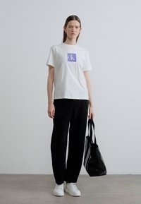 Άσπρο βαμβακερό t-shirt με μοβ λογότυπο, σε συνδυασμό με μαύρο παντελόνι και άσπρα αθλητικά παπούτσια. Μια ασπρόμαυρη τσάντα ώμου με ρίγες ολοκληρώνει την εμφάνιση.