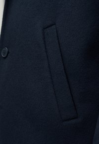 Détail d'un manteau en laine marine montrant une texture lisse avec une fermeture à boutons et une seule poche latérale avec un bord subtilement cousu.
