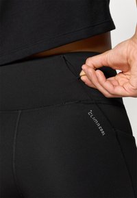 Crne sportske tajice s visokim strukom, mekom teksturom i kontrastnim šavovima. Imaju mali džep s patentnim zatvaračem straga.