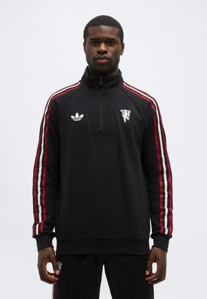 Άνδρας που φοράει μαύρο φούτερ με φερμουάρ έως το στήθος, με κόκκινες και λευκές ρίγες στα μανίκια, με λογότυπα Adidas και Manchester United στο στήθος, στέκεται όρθιος.