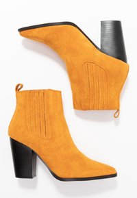 Bottes chevilles en daim jaune avec un bout pointu, des accents côtelés sur les côtés, et un talon noir épais. Languette de tirage visible à l'arrière.