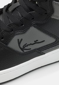 Zapatilla alta de cuero negro texturizado con acentos grises, que incluye cordones negros y una firma en negro en el lateral. Suela de goma blanca.