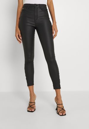 Femme portant un pantalon skinny en similicuir noir taille haute et des sandales à talons hauts noires à brides, debout sur un fond blanc.