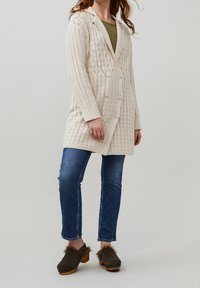 Cardigan en tricot beige clair avec un motif en losanges, équipé d'une capuche, de six boutons et de poignets côtelés. Associé à un jean bleu et des sabots marron.