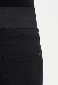 Närbild av svarta byxor med elastiskt midjeband och synlig söm på bakfickan samt nitar på en ren vit bakgrund.