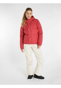 Rode pufferjack met capuchon, knoopsluitingen en zijzakken, gecombineerd met crèmekleurige broek en zwarte sneeuwlaarzen.