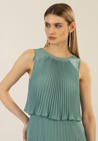 Ein plissiertes, ärmelloses Kleid aus zartem Teal-Stoff, mit einem runden Ausschnitt und einer fließenden Silhouette, die einen geschichteten Effekt aufweist.