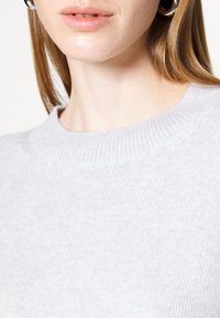 Pull en maille gris clair avec un col montant côtelé. Texture douce avec une finition lisse. Vue en gros plan de la zone du col.