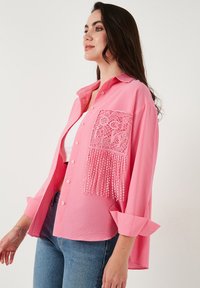Pink button-up shirt with textured fabric, featuring a lace pocket with fringe detail. Cuffed sleeves and a relaxed fit.