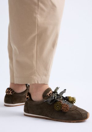 Zapatillas de ante marrón con detalles de estampado de leopardo y pompón decorativo, usadas con pantalones cortos beiges.