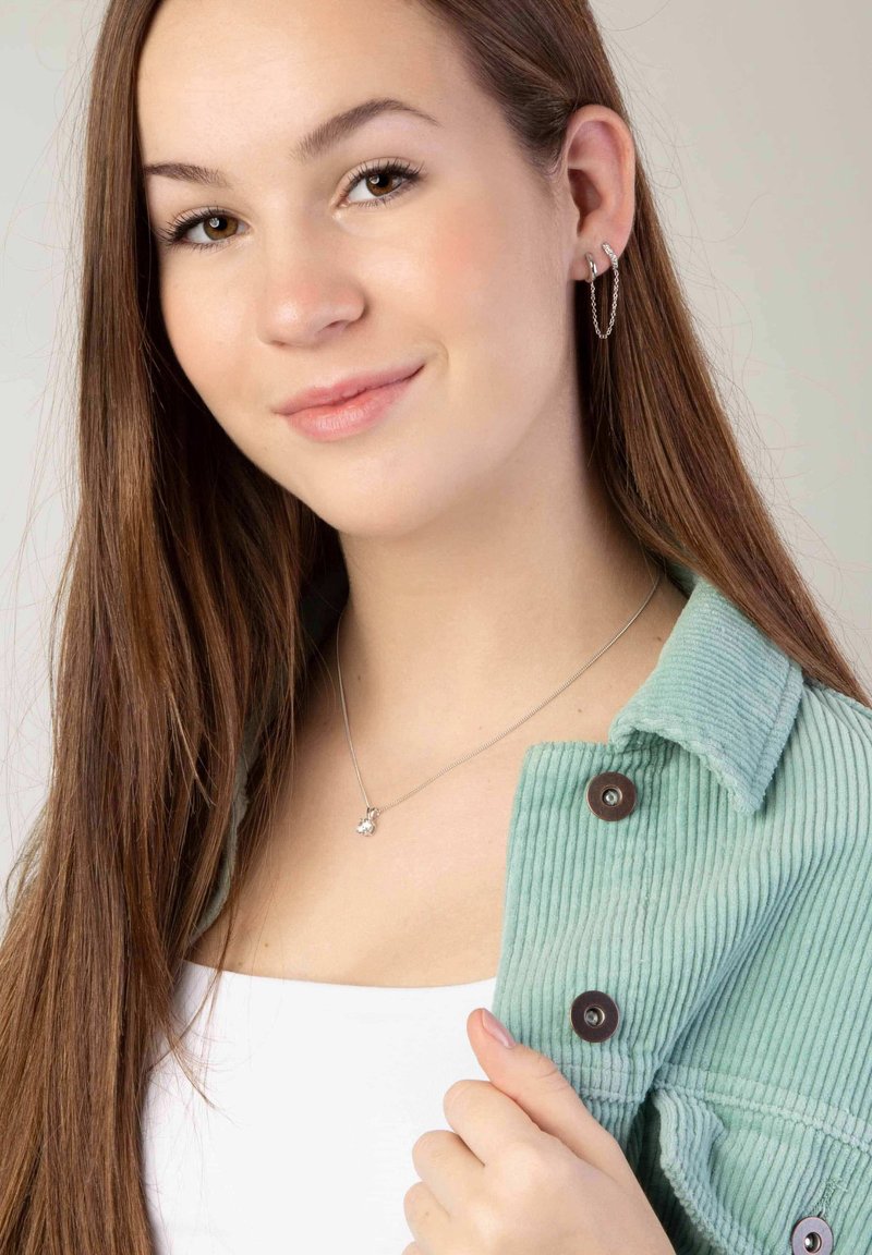 Veste en velours côtelé vert clair avec des accents de boutons, haut blanc et collier en argent avec un petit pendentif. Cheveux longs et bruns partiellement visibles.