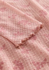 Světlé růžové krajkové tkaniny s texturovaným květinovým vzorem a zvlněnými okraji. Materiál vypadá měkký a jemný.
