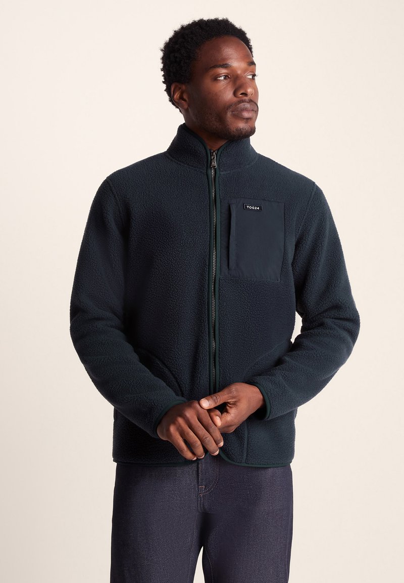 Dark green fleece jacket with a high collar, zip closure, and a front chest pocket. Soft texture and contrasting pocket fabric detail.