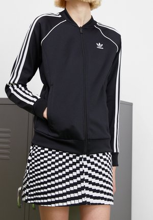 Person iført en sort Adidas lynlåstrøje med hvide striber på ærmerne og et sort-hvidt nederdel med geometrisk mønster.