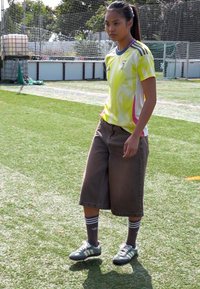 Idrottare i en gul och vit kortärmad tröja med rosa detaljer, bruna vida shorts, grå fotbollsskor och randiga strumpor.