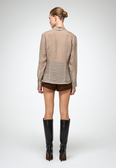 Camicia beige semi-trasparente a maniche lunghe con colletto, polsini abbottonati e vestibilità rilassata, abbinata a pantaloni corti marroni scuri e stivali neri alti.