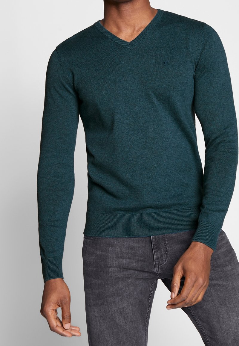 Homme portant un pull à col en V manches longues couleur sarcelle et un jean gris foncé, debout devant un fond clair uni.