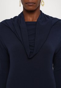 Námorně modrý dlouhý rukáv s draped cowl výstřihem. Vyrobeno z jemné tkaniny, s jemnou strukturou a přiléhavým střihem.