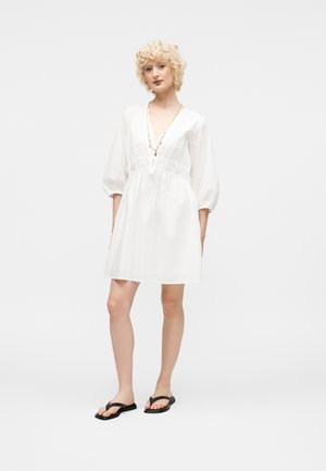 Młoda osoba z krótkimi kręconymi blond włosami, ubrana w białą sukienkę do kolan z rękawami do łokcia i czarne płaskie sandały.