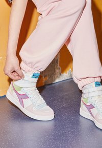 Person justerar vita högsnörade sneakers med pastellfärger i rosa, lila, grön och blå, klädd i rosa joggingbyxor på ett prickigt blått golv.